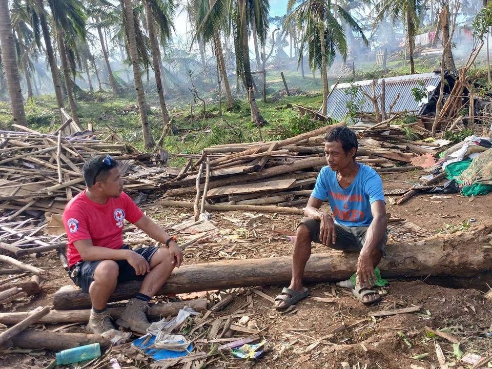 A Red Cross volunteer speaks to a man in the Philippines infront of a destroyed house with palm trees in the background.