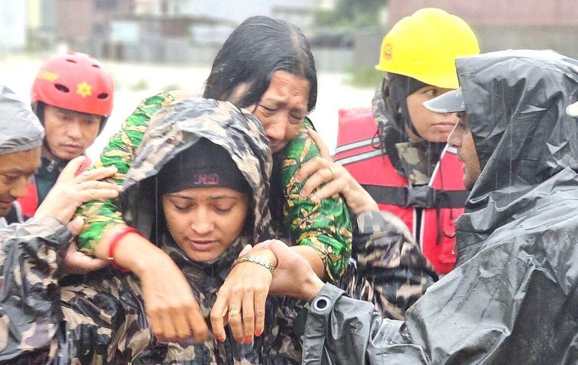 A women in Nepal is carried on the back of another women in the rain. There are people in the background and everyone wears rainjackets.