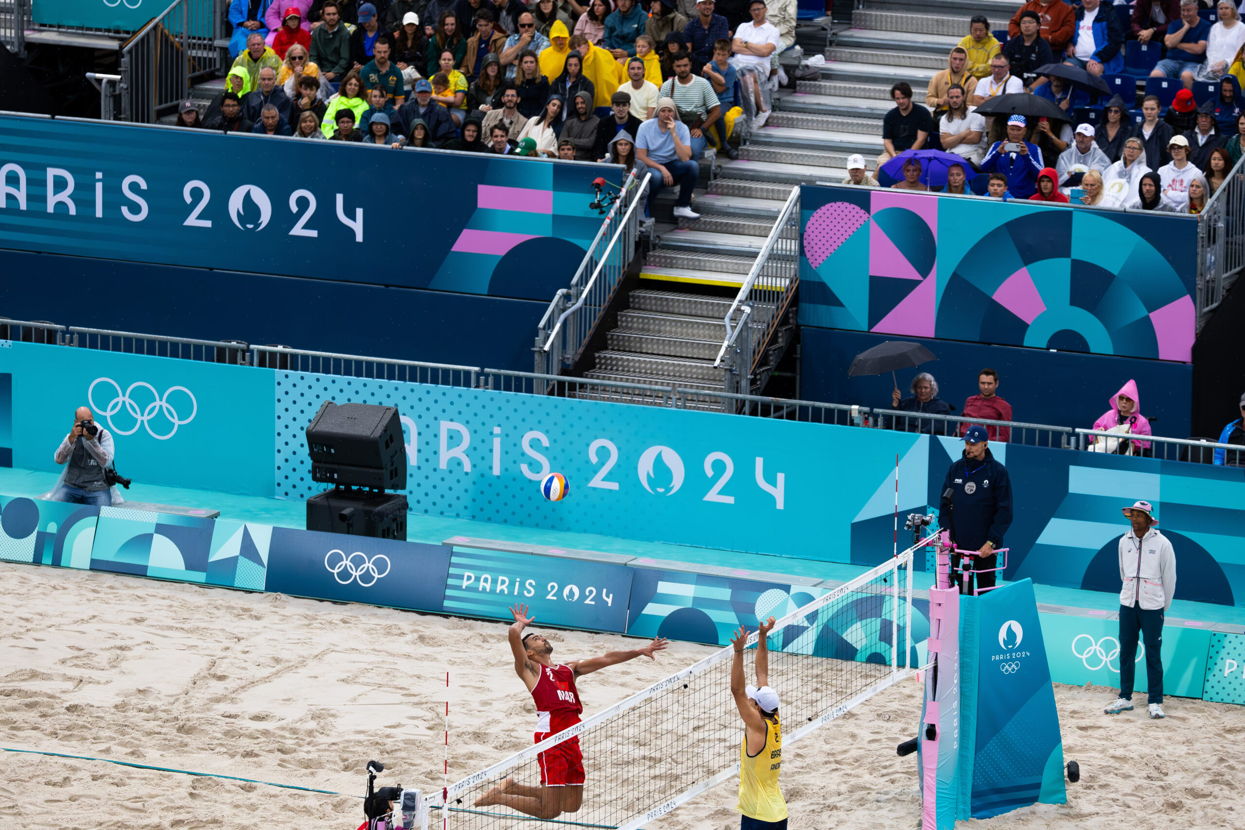 Mens beach volleyball at the Olympics. A player in red leaps into the air preparing to hit the ball. Another player in yellow on the other side of the year jumps with his hands in the air to block the oncoming shot. Photographers and a crowd on a stand are in the background.