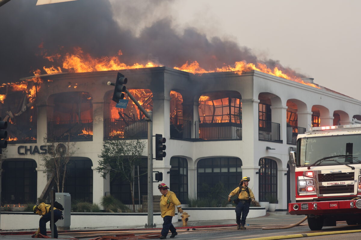 Two firefighters stand infront of a burning banlk in LA. It is a two story white building with large flames burning from the roof.