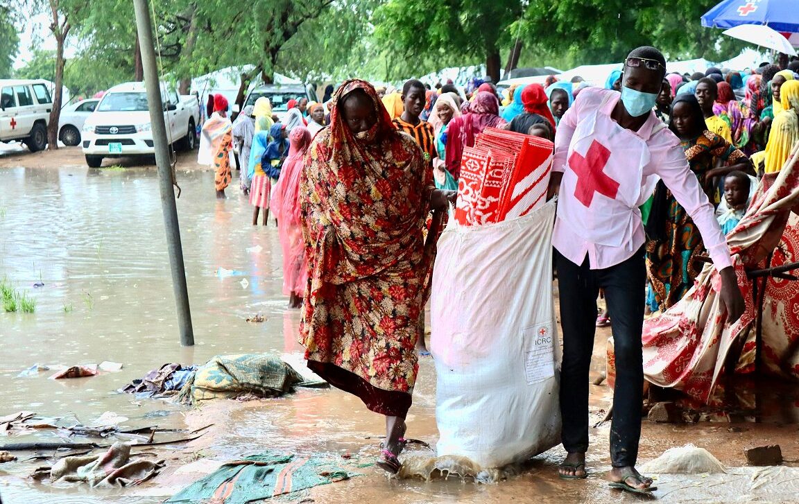 A man in a white red cross bib assists a woman walking next to floodwaters in Chad.