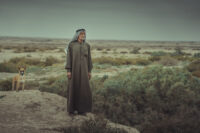 A photo of a man in Iraq, wearing traditional clothing, standing next to a dog and in front of a dried up river.